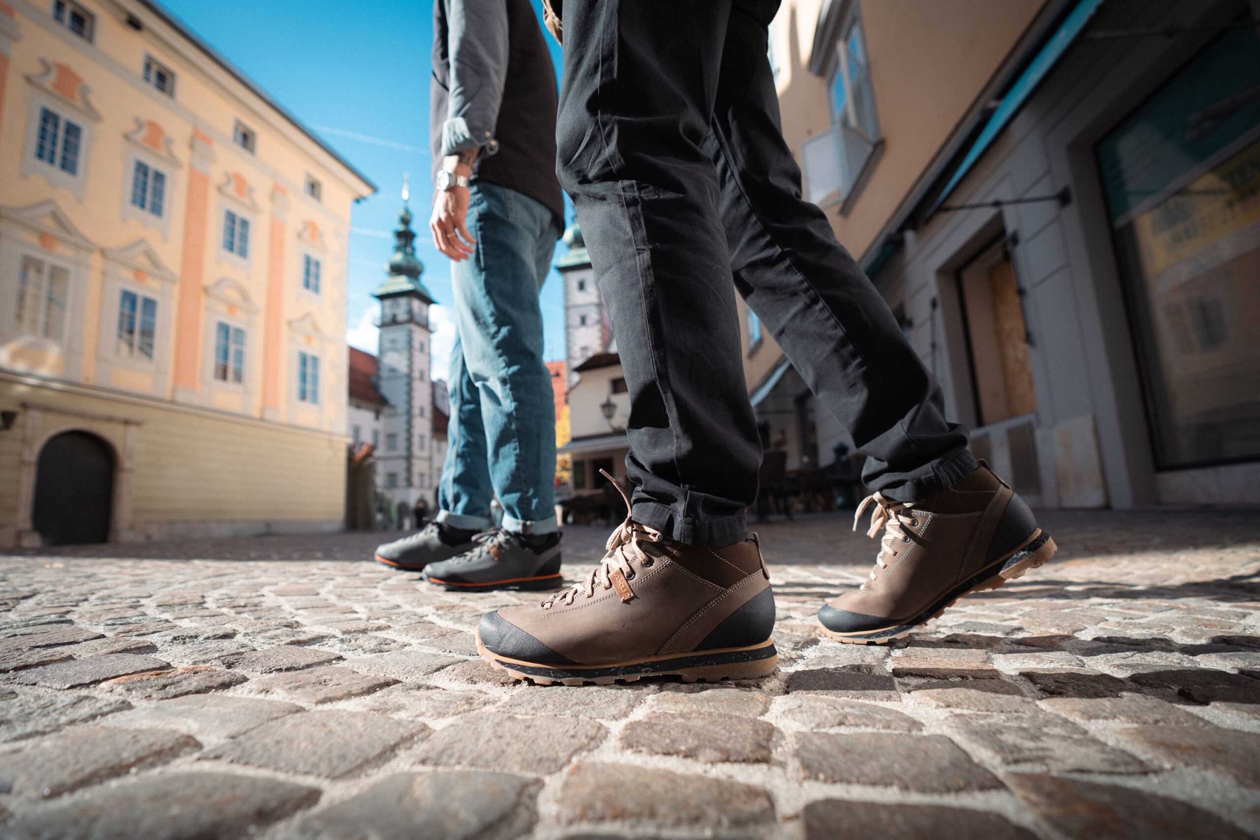Two people walking on a cobblestone street in a sunlit town square.