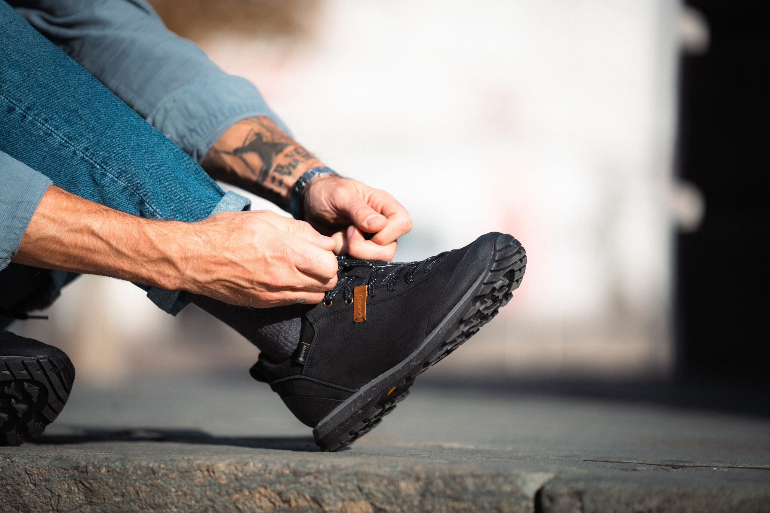 Person with tattooed arm tying black hiking boots while sitting outdoors.